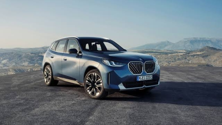 Vista frontal del SUV BMW X3 premium estacionado junto a una costa con montañas de fondo.